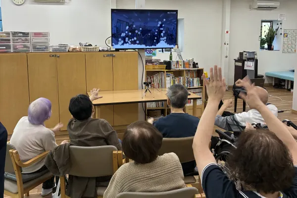 デイサービスで、テレビに映ったゲームに合わせて、高齢者が椅子に座ったまま手を上げている写真